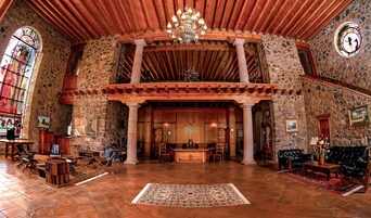 A spacious lobby with a high ceiling, stone walls, and a wooden ceiling. There is a chandelier, a reception desk, and a large rug on the floor.