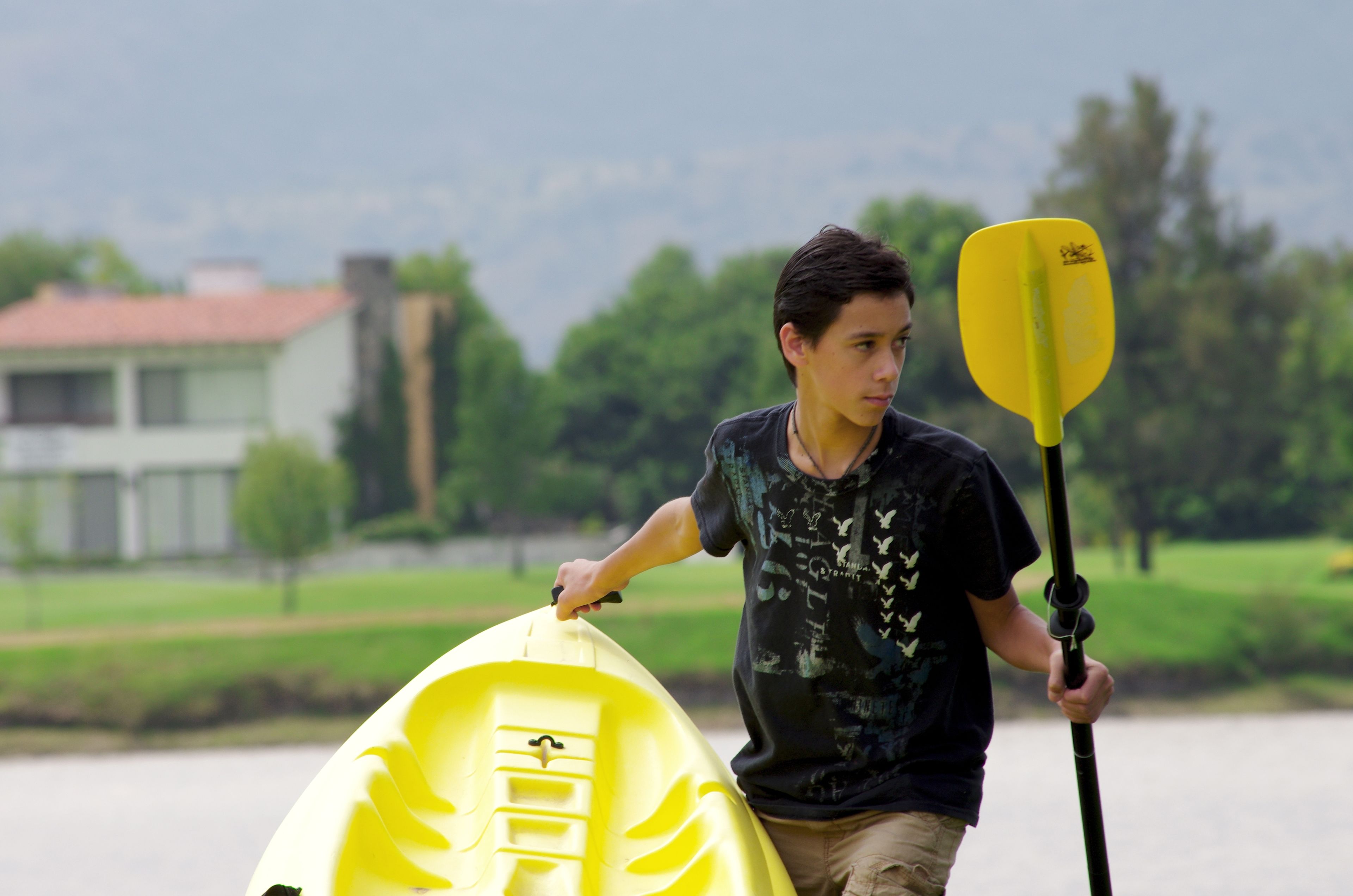 A person holding a yellow kayak and a paddle, standing by a body of water with a house and greenery in the background.
