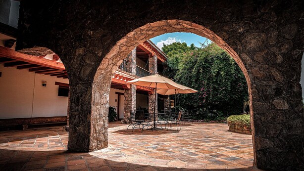 Un patio al aire libre con pilares de piedra, una zona de estar cubierta y vistas a un edificio con tejado rojo.