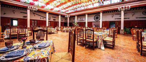 Un comedor amplio con techos altos decorados con coloridos adornos de papel, mesas y sillas de madera, y suelo de ladrillo.