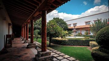 A covered walkway with stone columns, a garden with a fountain, and a building with a tiled roof.