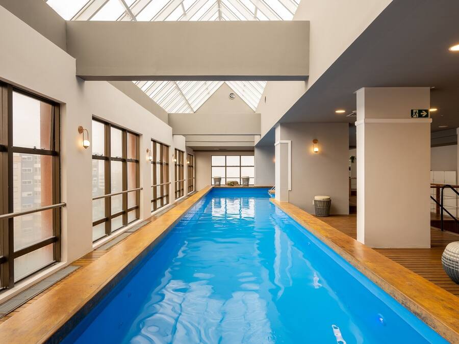 A long indoor swimming pool with wooden deck edges, large windows on one side, and a skylight above.