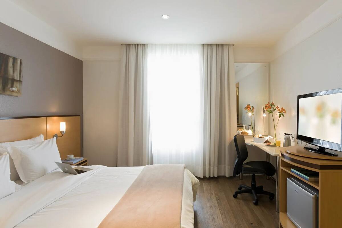 Une chambre d’hôtel équipée d’un lit, d’un bureau avec un ordinateur, d’une chaise, d’une télévision et d’un petit réfrigérateur.