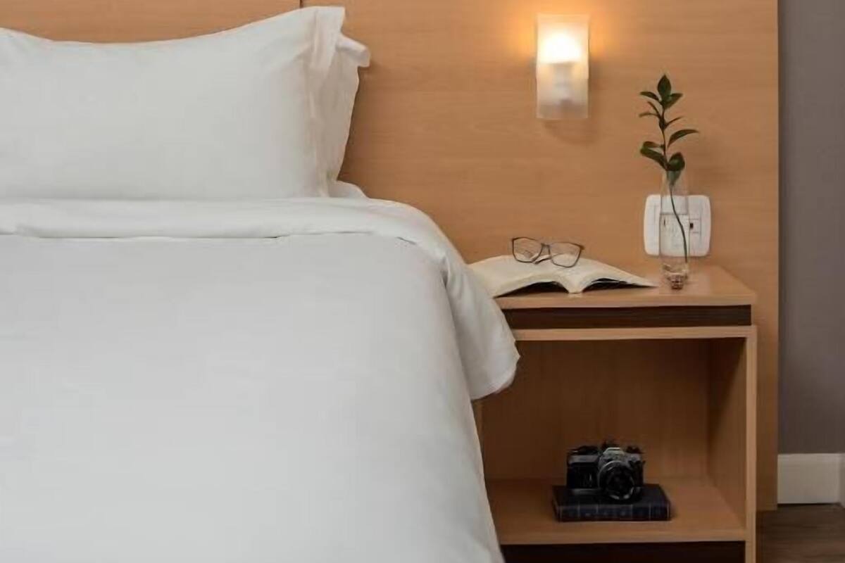 Un lit bien fait, avec du linge de lit blanc, une tête de lit en bois, une table de chevet avec une lampe, une petite plante et un appareil photo.