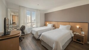 A hotel room with two beds, a desk with a computer, a chair, a nightstand with a phone and a plant, and a view of the city through large windows. - Intercity Caxias do Sul (Caxias do Sul)