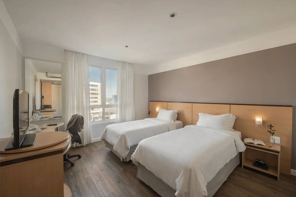 Une chambre d’hôtel avec deux lits, un bureau avec un ordinateur, une chaise, une table de chevet avec un téléphone et une plante, et une vue sur la ville grâce à de grandes fenêtres.