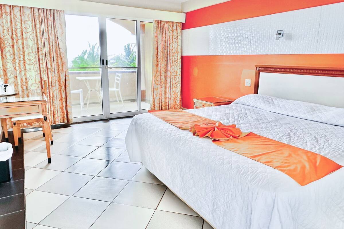 A hotel room with a large bed, a desk, and a chair. There is a sliding glass door leading to an outdoor area.