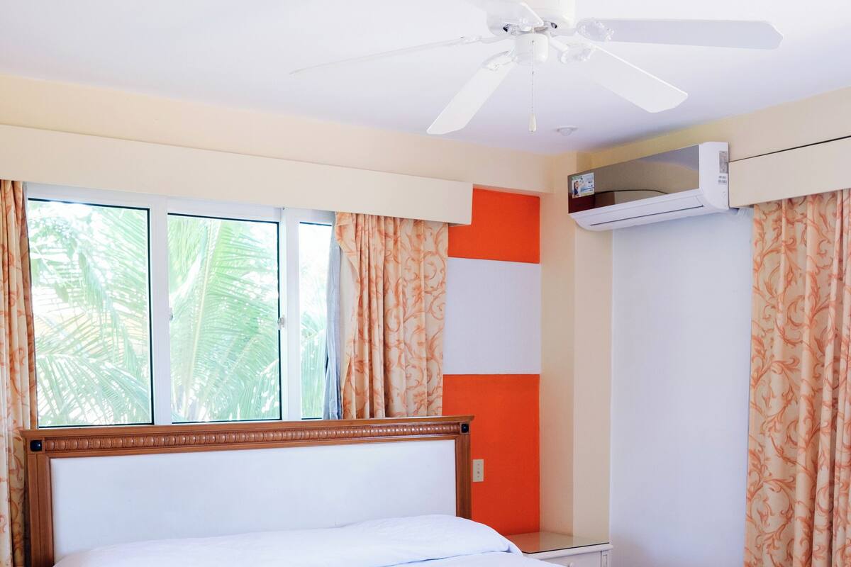 A hotel room with a bed, a ceiling fan, an air conditioning unit, and curtains.