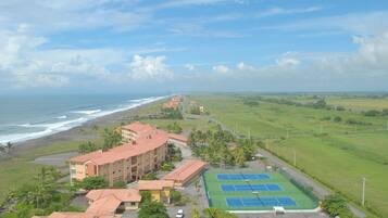 A coastal hotel with a tennis court, beachfront view, and multiple buildings.