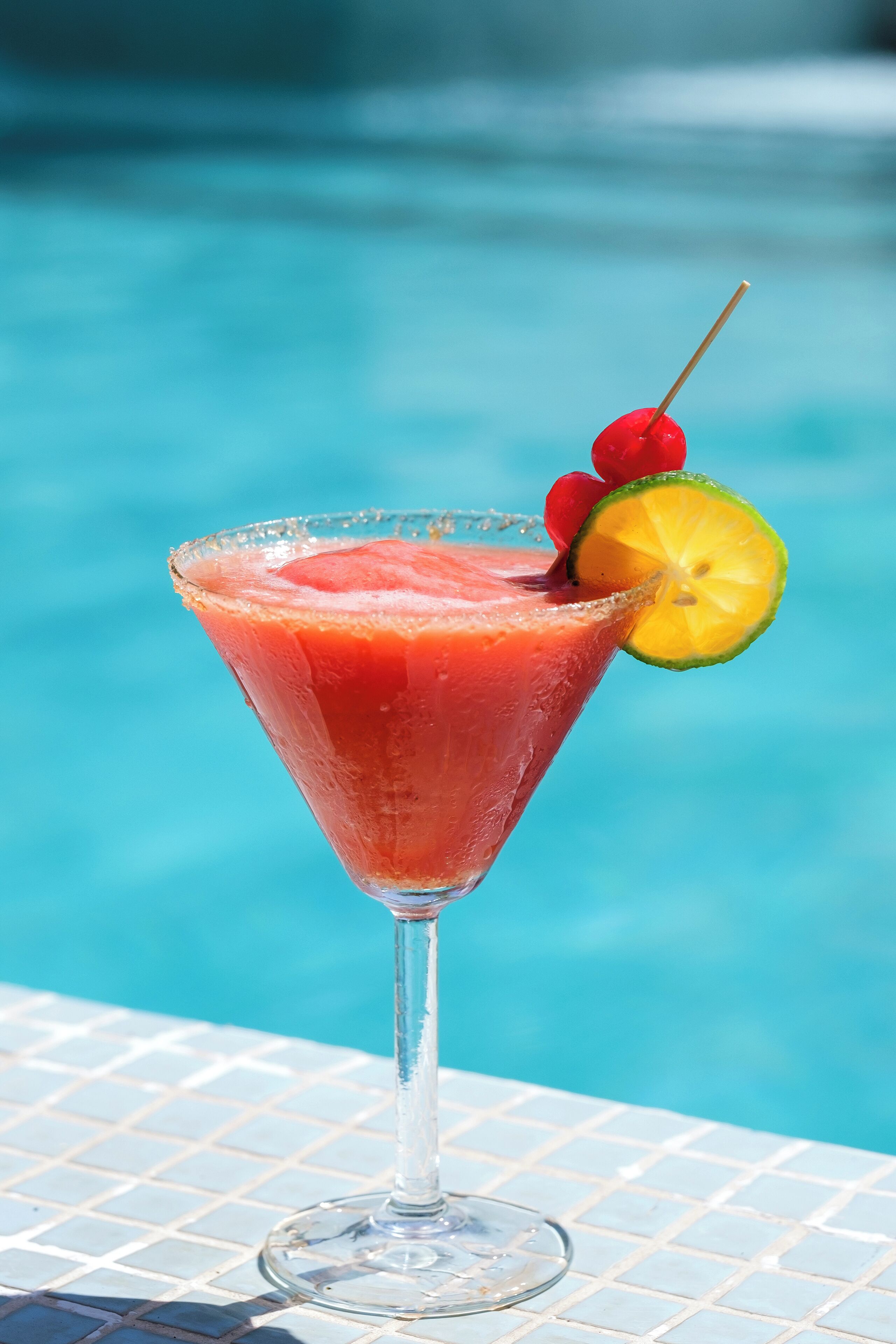 A cocktail glass with a slice of lime and a cherry on a tiled poolside table.