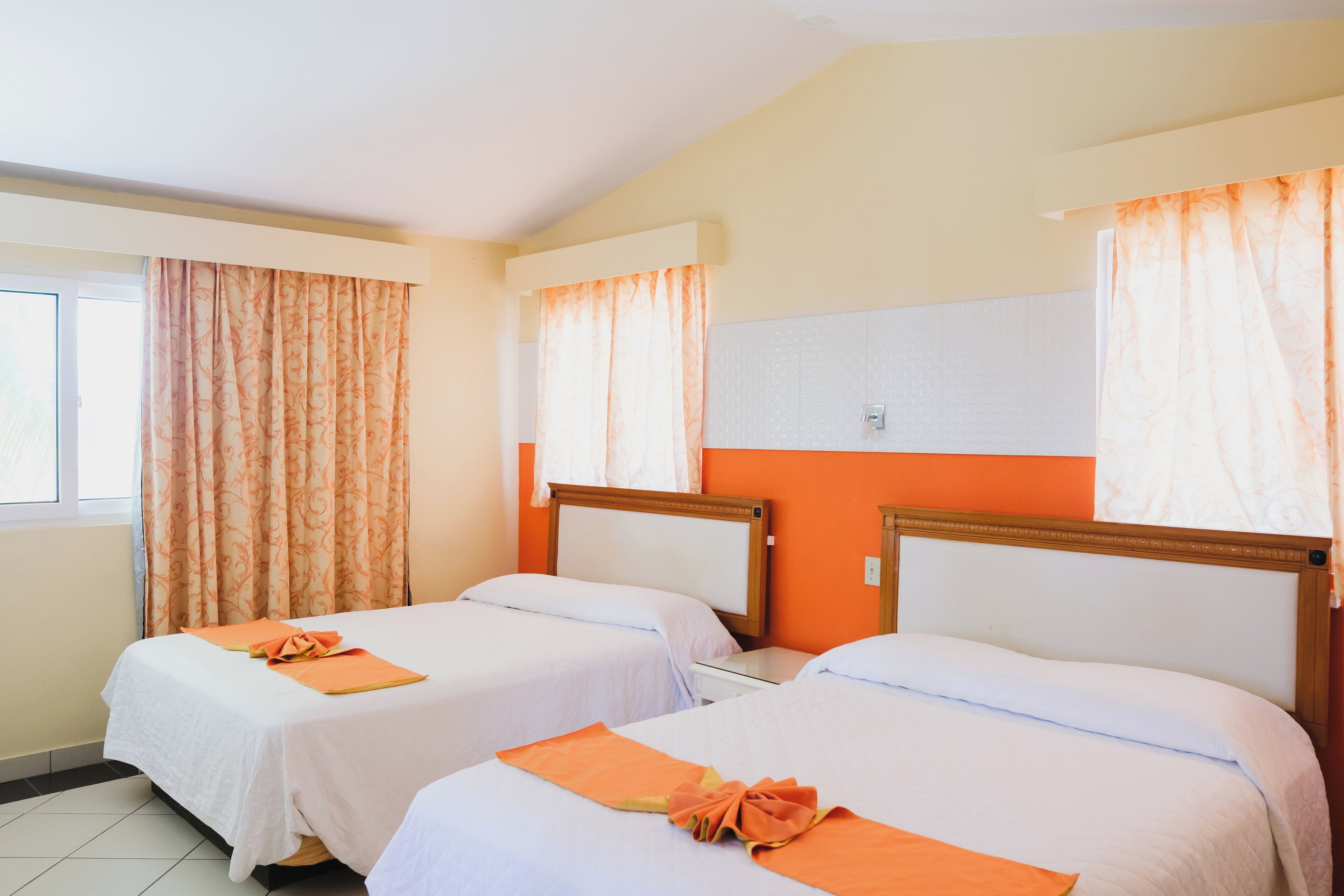 Quarto de hotel com duas camas de solteiro, cada uma com uma toalha laranja e roupa de cama branca. O quarto tem paredes laranja e uma janela com cortinas estampadas.