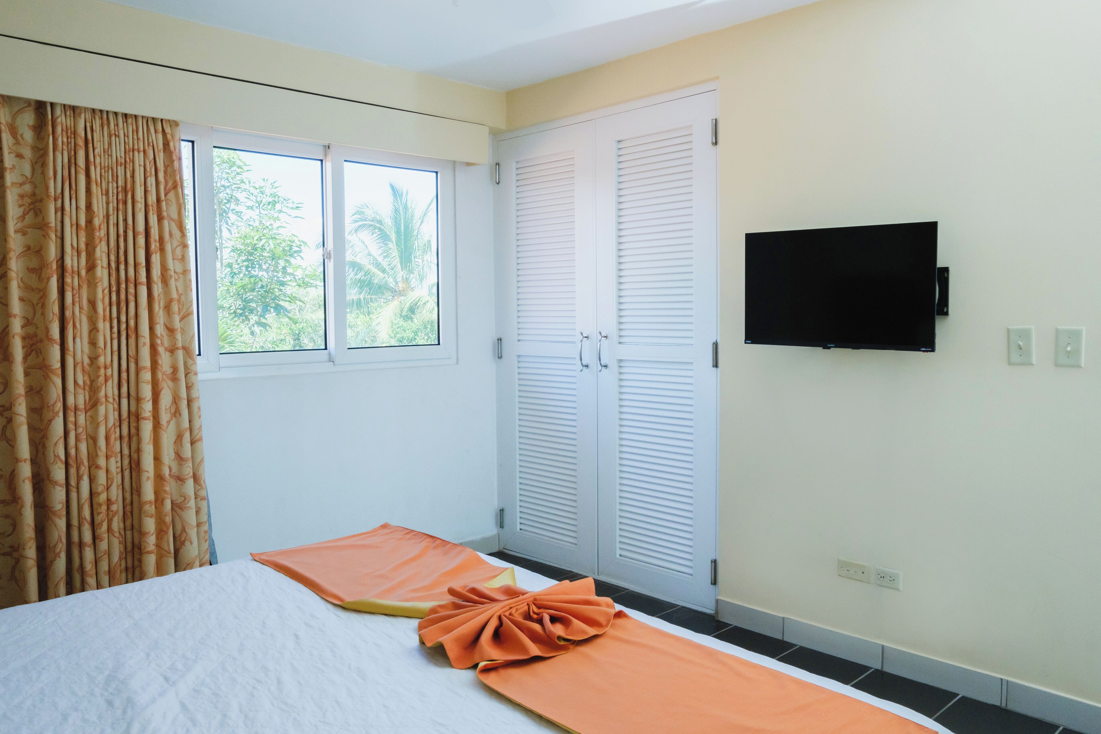 Un dormitorio con cama, cortinas, televisor y ventana con vista a follaje.