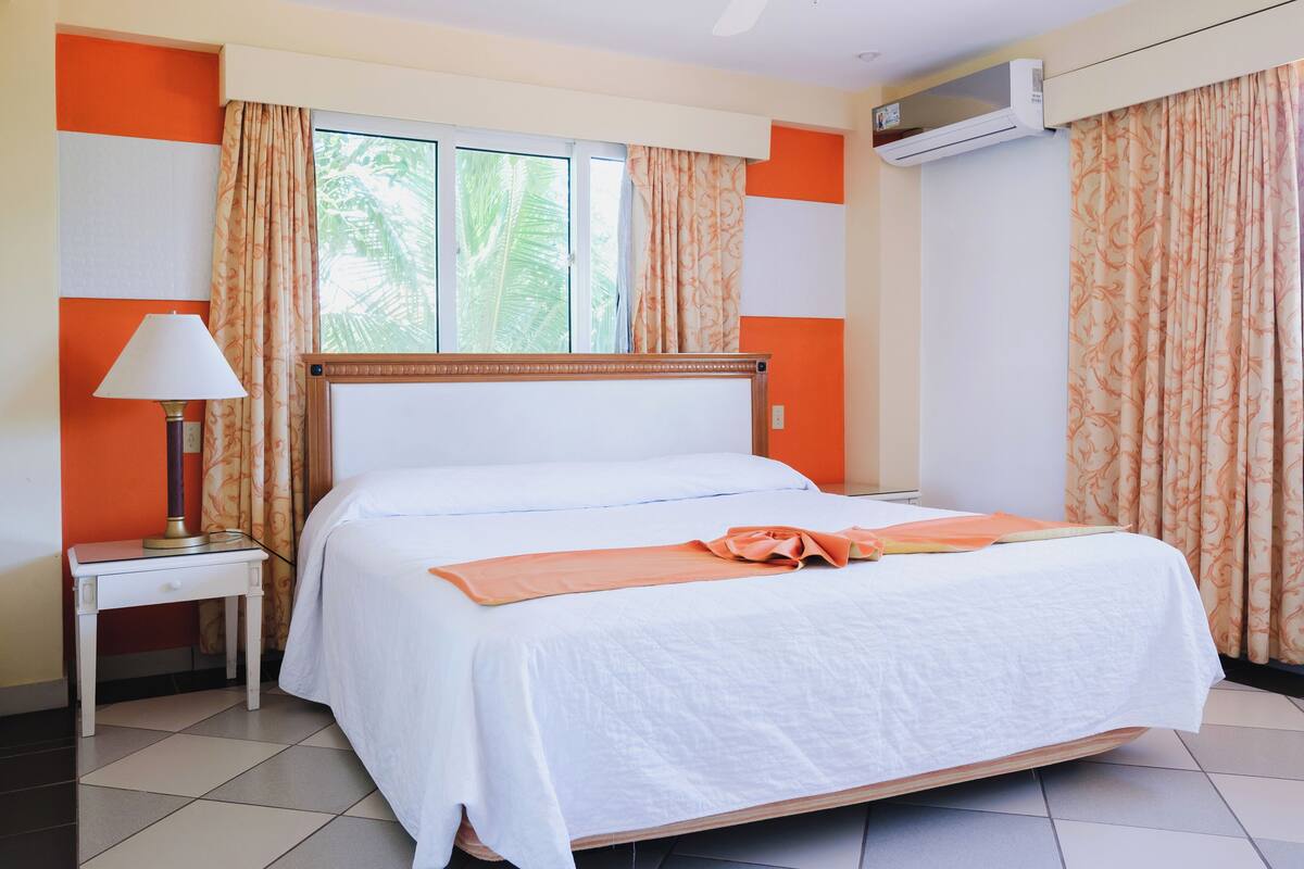A hotel room with a bed, a nightstand, an air conditioning unit, and curtains.