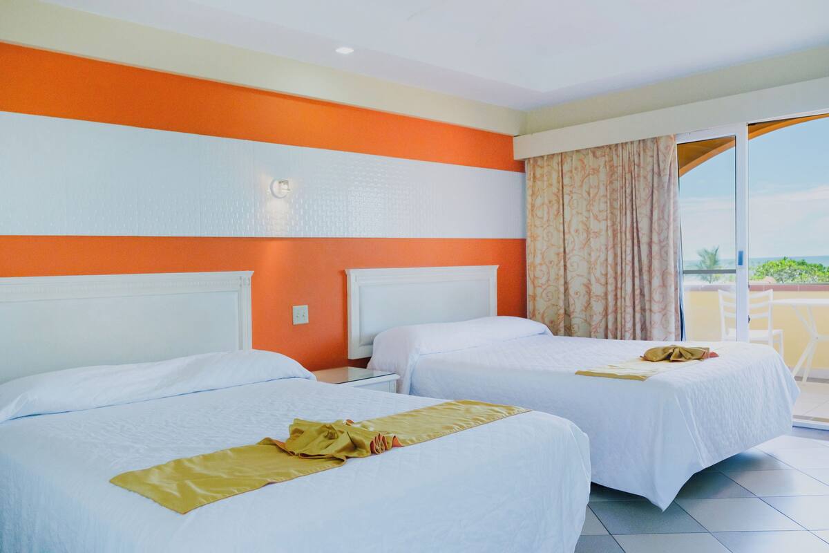 A hotel room with two beds, orange and white walls, a balcony with a view, and a window with curtains.