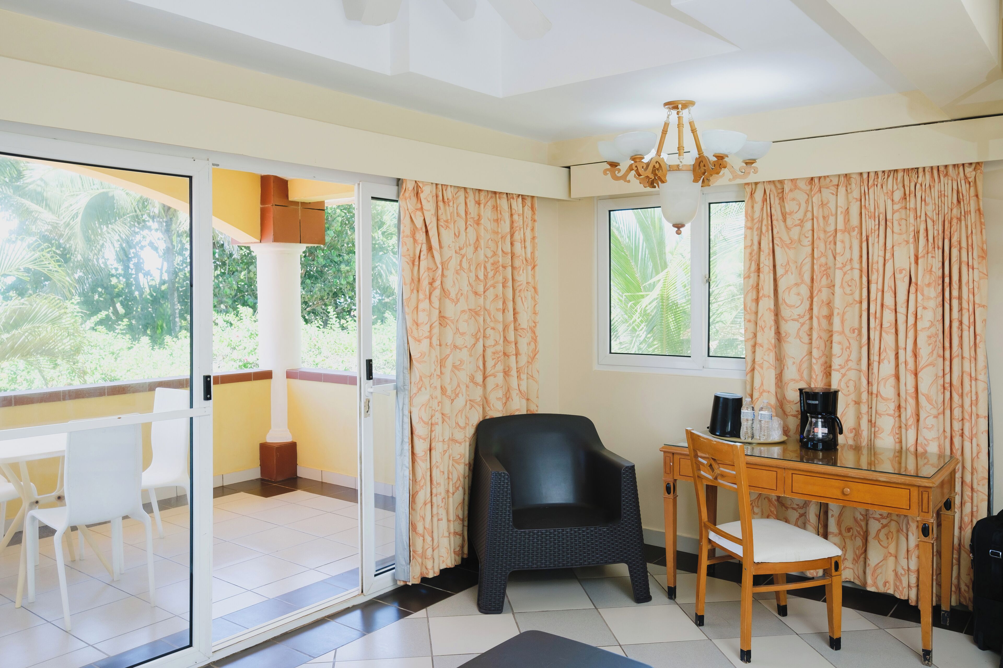 A room with a glass door leading to a balcony, a wooden desk with a chair, a black chair, and a coffee maker.