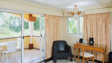 A room with a glass door leading to a balcony, a wooden desk with a chair, a black chair, and a coffee maker.