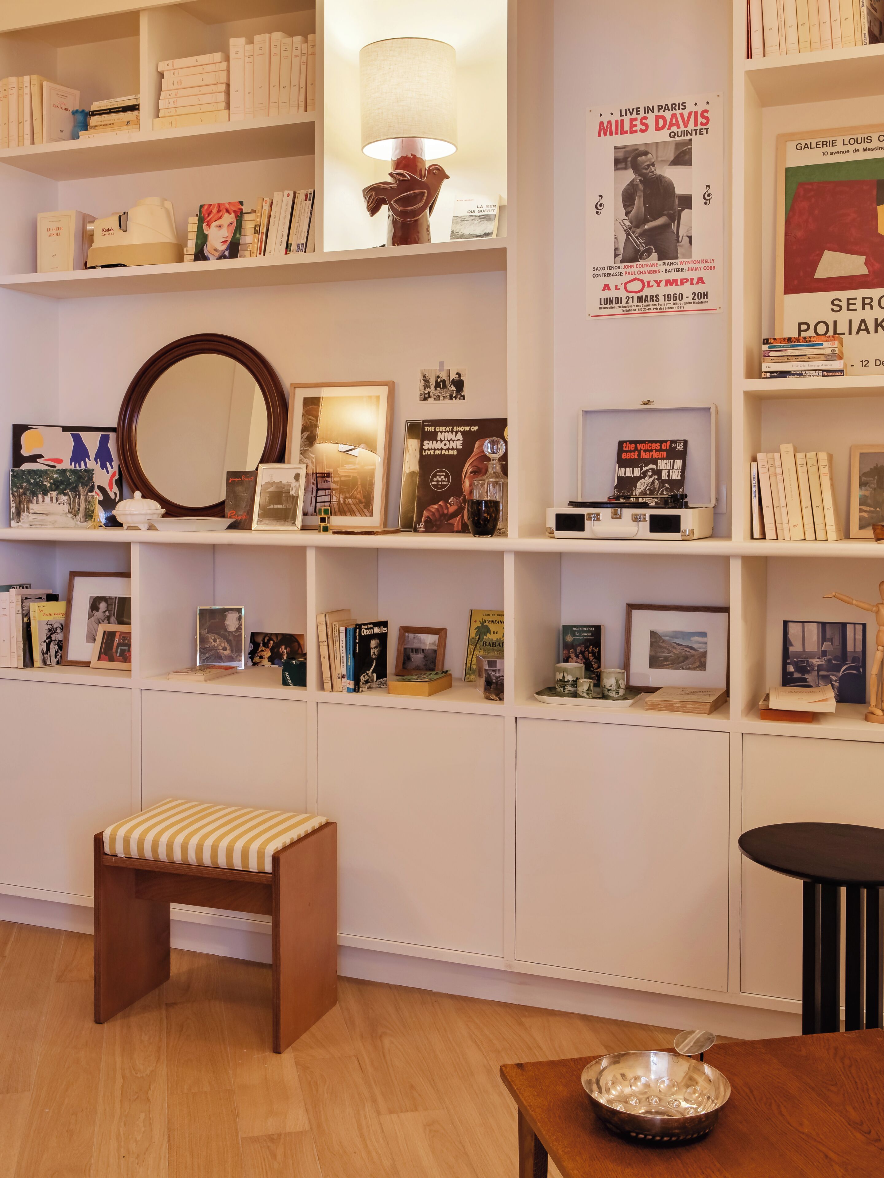 Une pièce avec un plancher en bois, un banc blanc, une petite table avec un bol, et des étagères remplies de livres et de photos encadrées.
