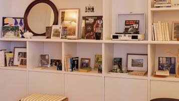 Une pièce avec un plancher en bois, un banc blanc, une petite table avec un bol, et des étagères remplies de livres et de photos encadrées.