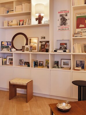 A room with a wooden floor, a white bench, a small table with a bowl, and shelves with books and framed pictures.