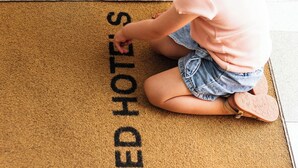 Ein Kind sitzt auf einer Fußmatte mit dem Schriftzug 'ALFRED HOTELS'.