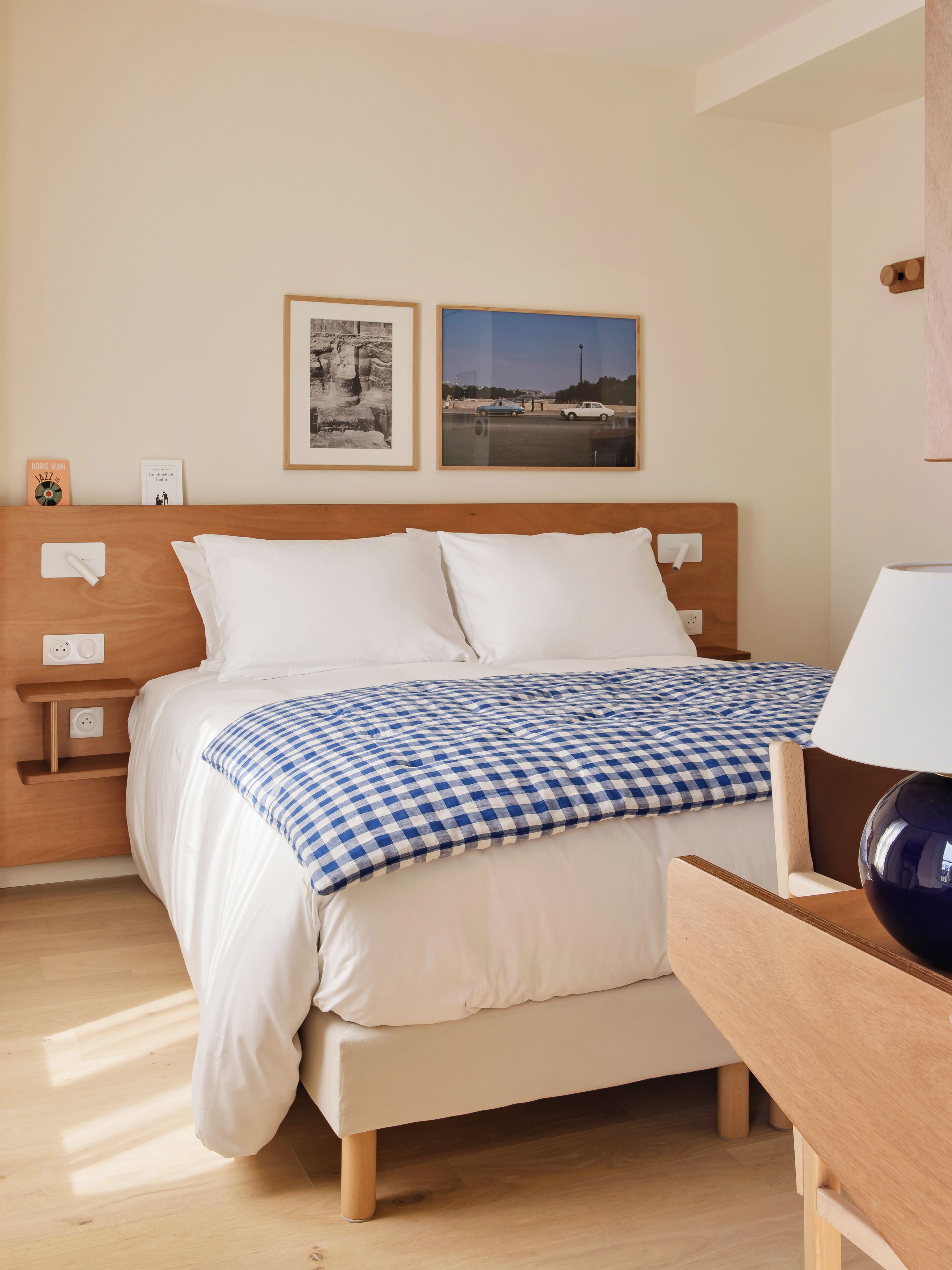 Una cama bien hecha con una colcha a cuadros, una mesita de noche con una lámpara y dos cuadros enmarcados en la pared.