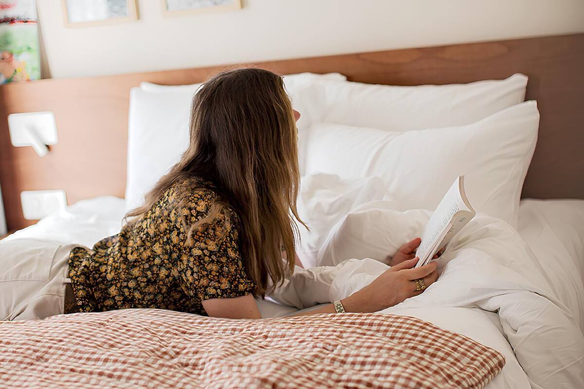 Une personne est allongée sur un lit, recouverte d’une couverture à carreaux, et lit un livre. La chambre est dotée d’une tête de lit en bois, de draps blancs et de deux tableaux encadrés accrochés au mur.
