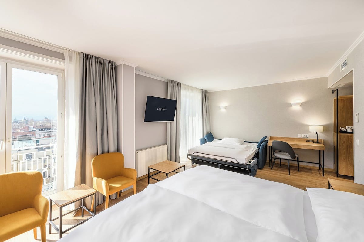 A hotel room with a bed, a desk, a chair, a TV, and a view of the city.