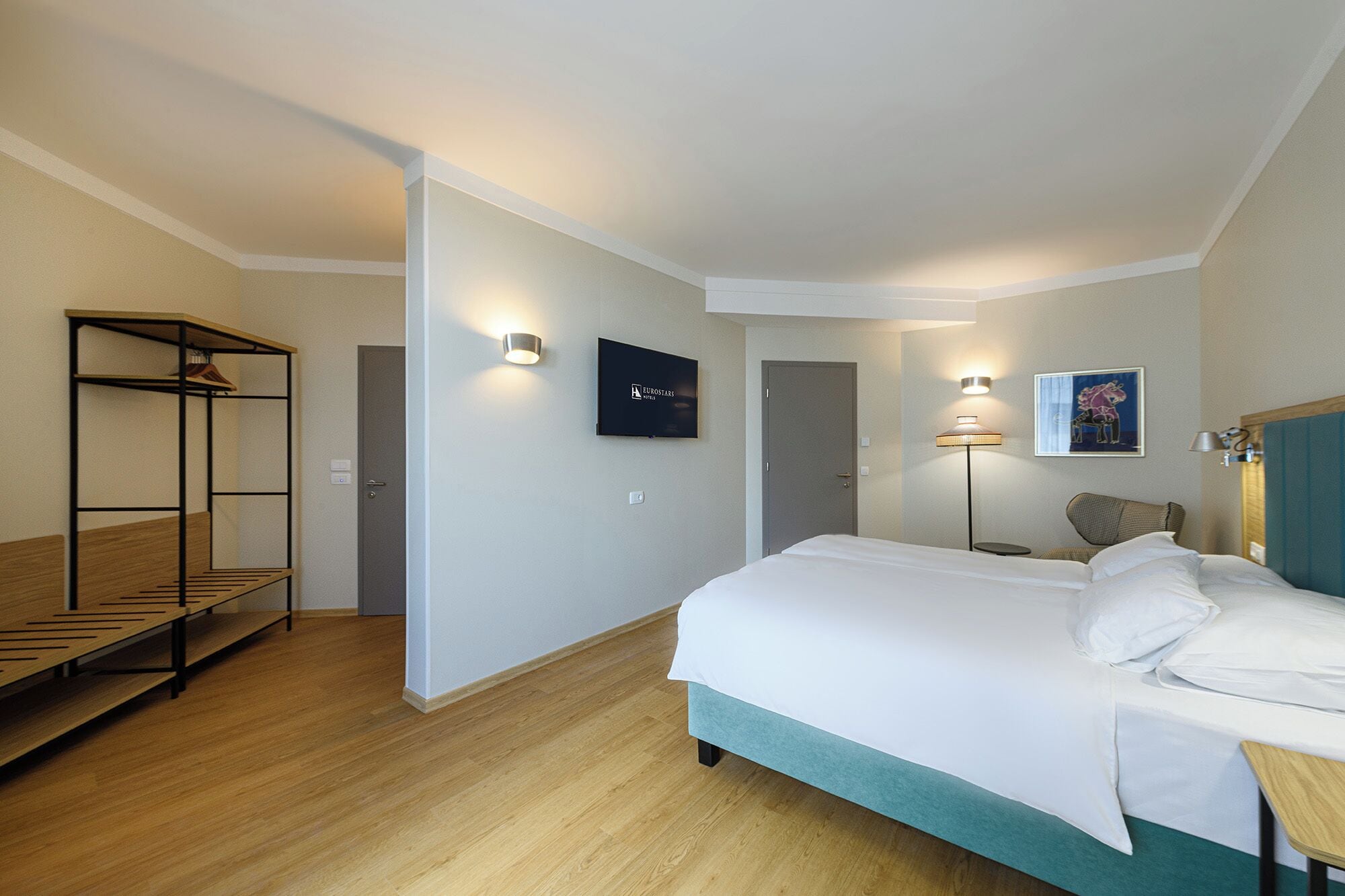 A hotel room with a bed, a seating area with a bench, a TV, a door, and a lamp.