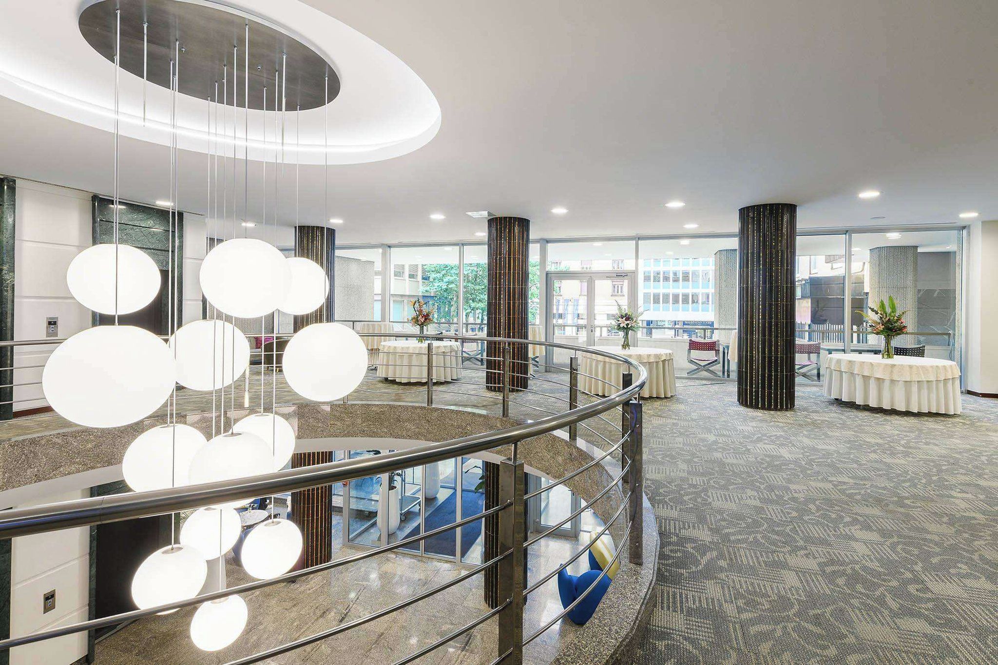 A modern hotel lobby with a central staircase, round tables, and a glass railing.