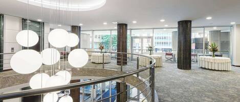 A modern hotel lobby with a central staircase, round tables, and a glass railing.
