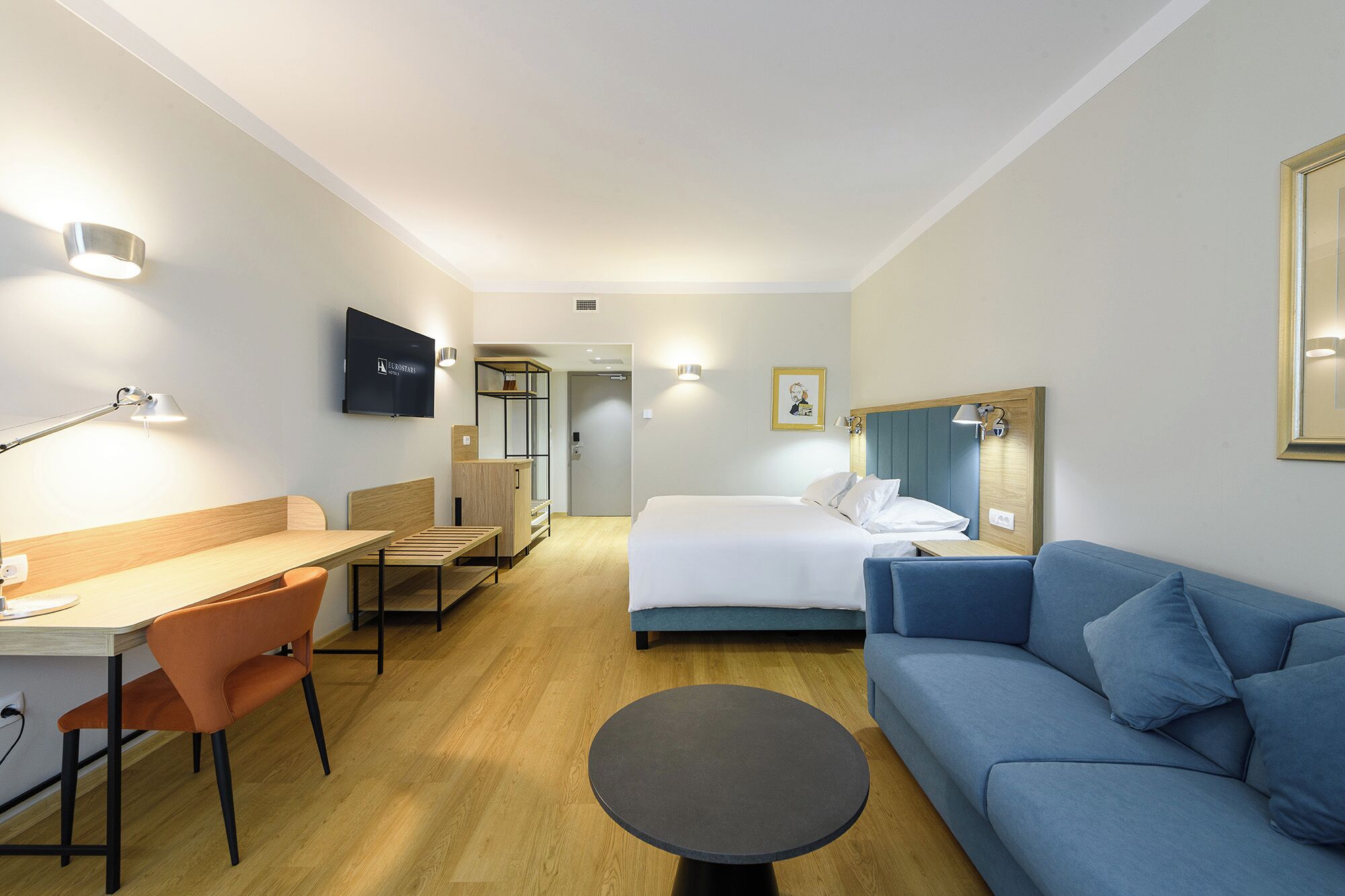 A hotel room with a bed, a desk with a chair, a sofa, a small table, and a TV on the wall.