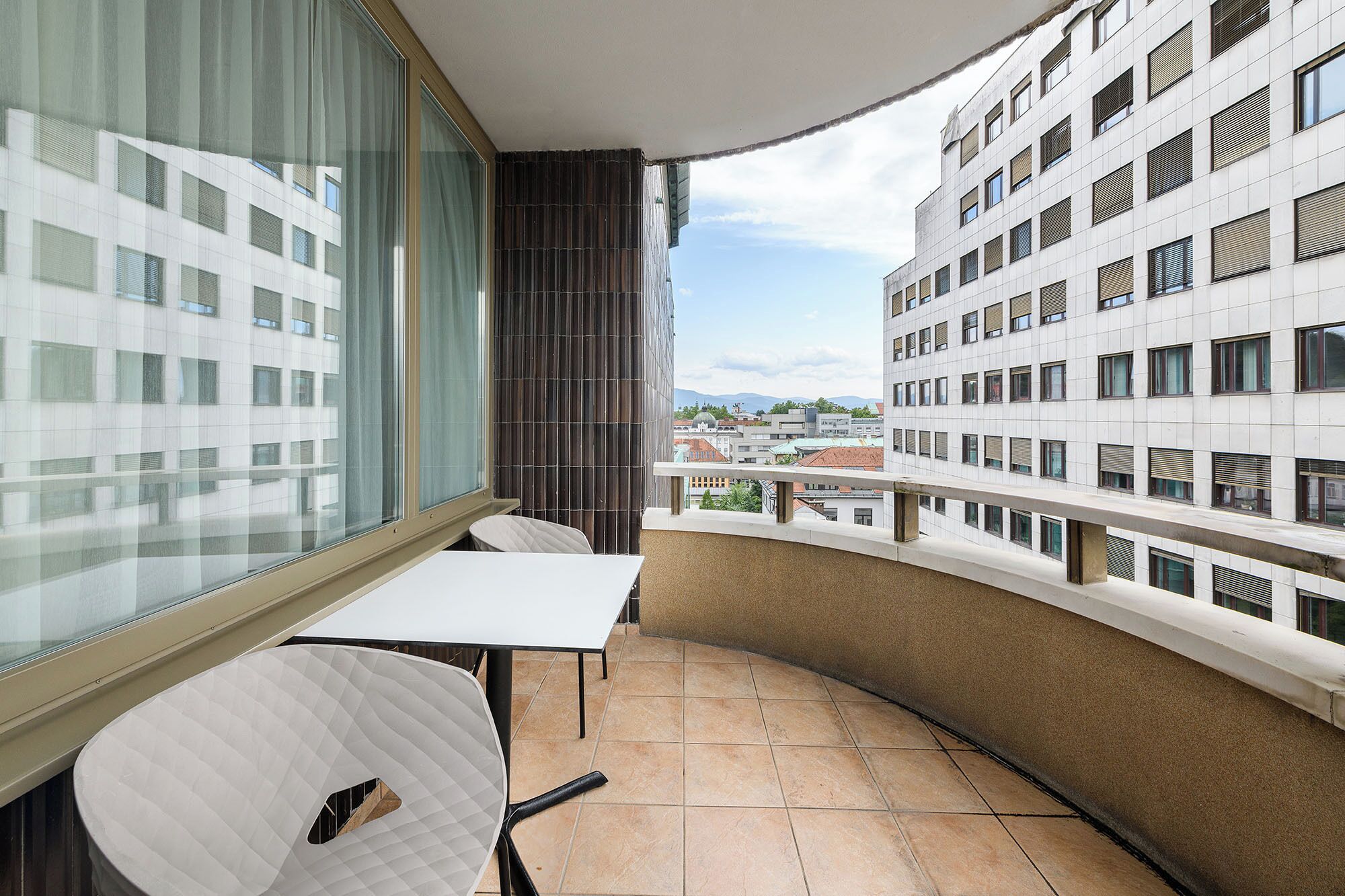 Un balcón con una mesa y sillas blancas, vista a un paisaje urbano y un edificio con fachada de diseño enrejado.