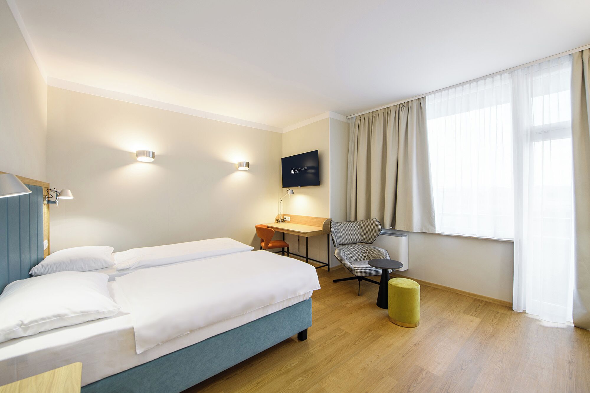 Ein Hotelzimmer mit einem Bett, einem Schreibtisch, einem Sessel, einem Fernseher und einem Fenster mit Vorhängen.