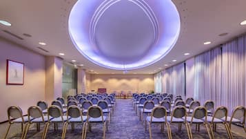 A conference room with rows of chairs, a stage, and a large circular light fixture on the ceiling.