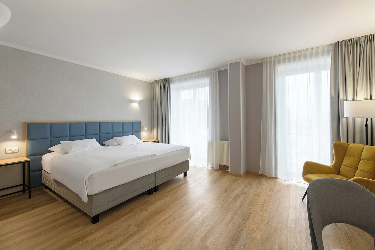 A modern hotel room with a large bed, a bedside table, a chair, and a lamp.
