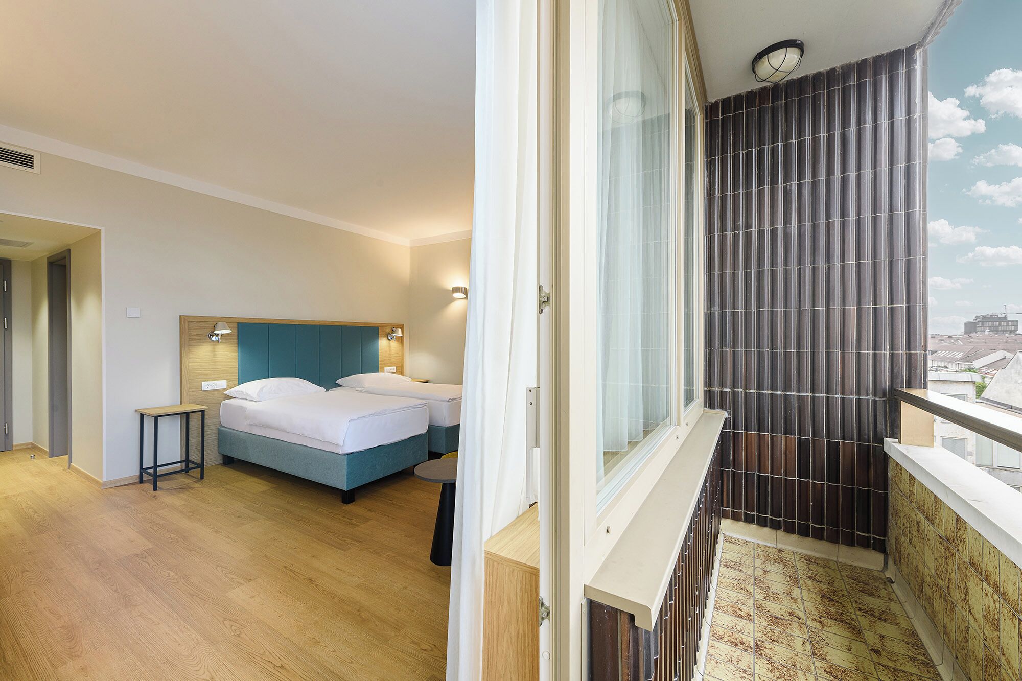 A hotel room with a bed, bedside table, and a view of the city through a window.