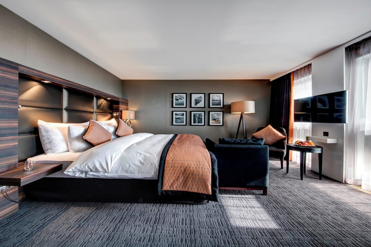 A modern hotel room with a large bed, a desk, a chair, a TV, and a window with curtains.