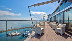 A sunlit deck with white chairs and a glass railing overlooking a marina.