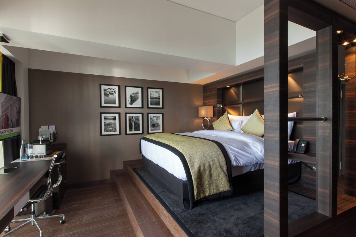 A modern hotel room with a bed, desk, and chairs. There are framed pictures on the wall and a TV on the desk.