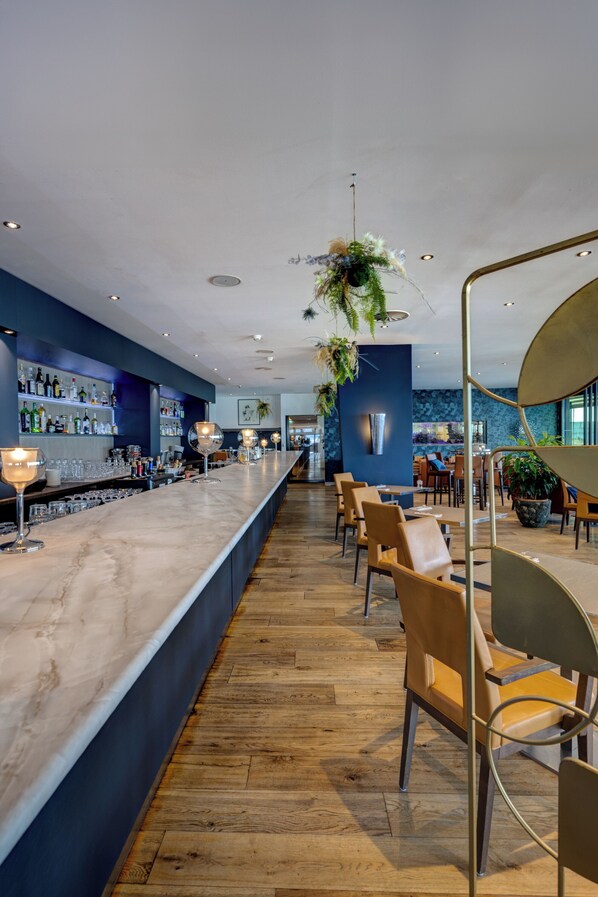 A bar area with a long counter, wooden flooring, and bar stools. There are shelves with bottles and glasses on the left.