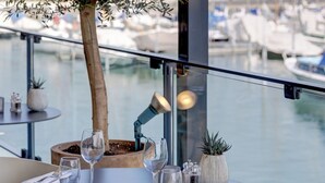 A table with a potted olive tree, wine glasses, and a view of a marina with boats.