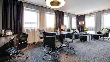 Quarto de hotel com mesa de conferência, cadeiras, sofá e escrivaninha com abajur.