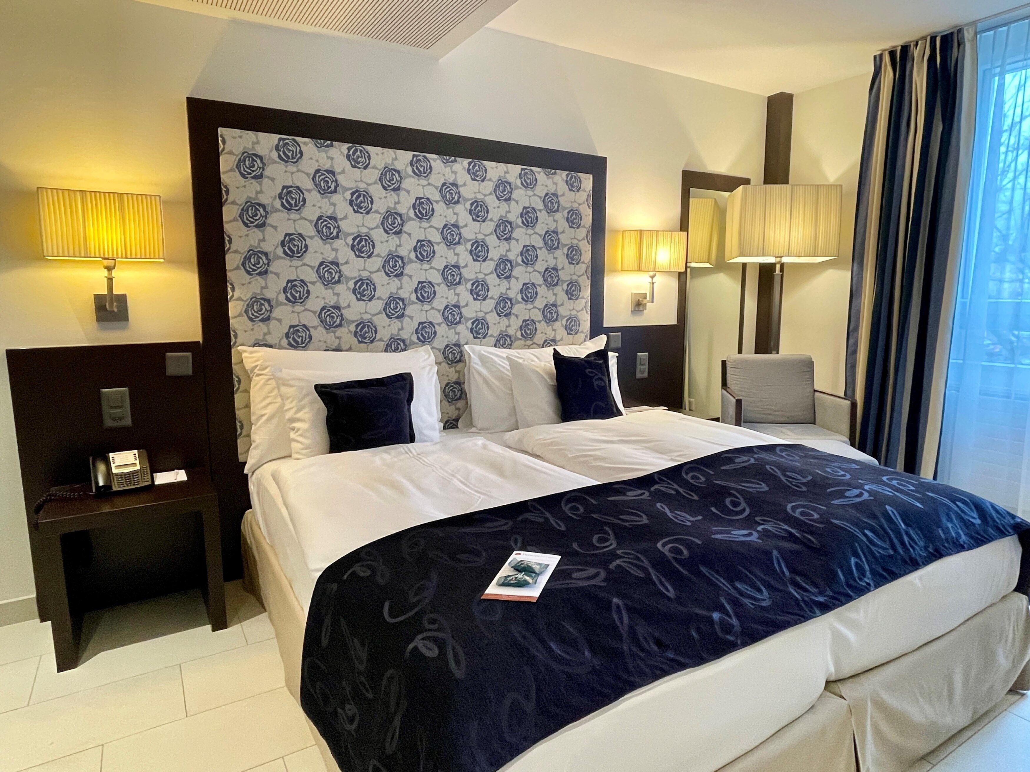 Una habitación de hotel con una cama grande, dos mesitas de noche, una silla y una ventana con cortinas.