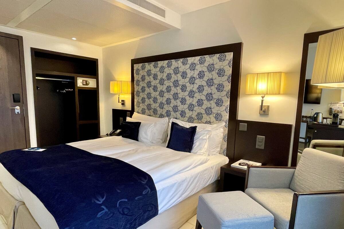 A hotel room with a large bed, a bedside table with a lamp, a chair, and a small table with a coffee maker.