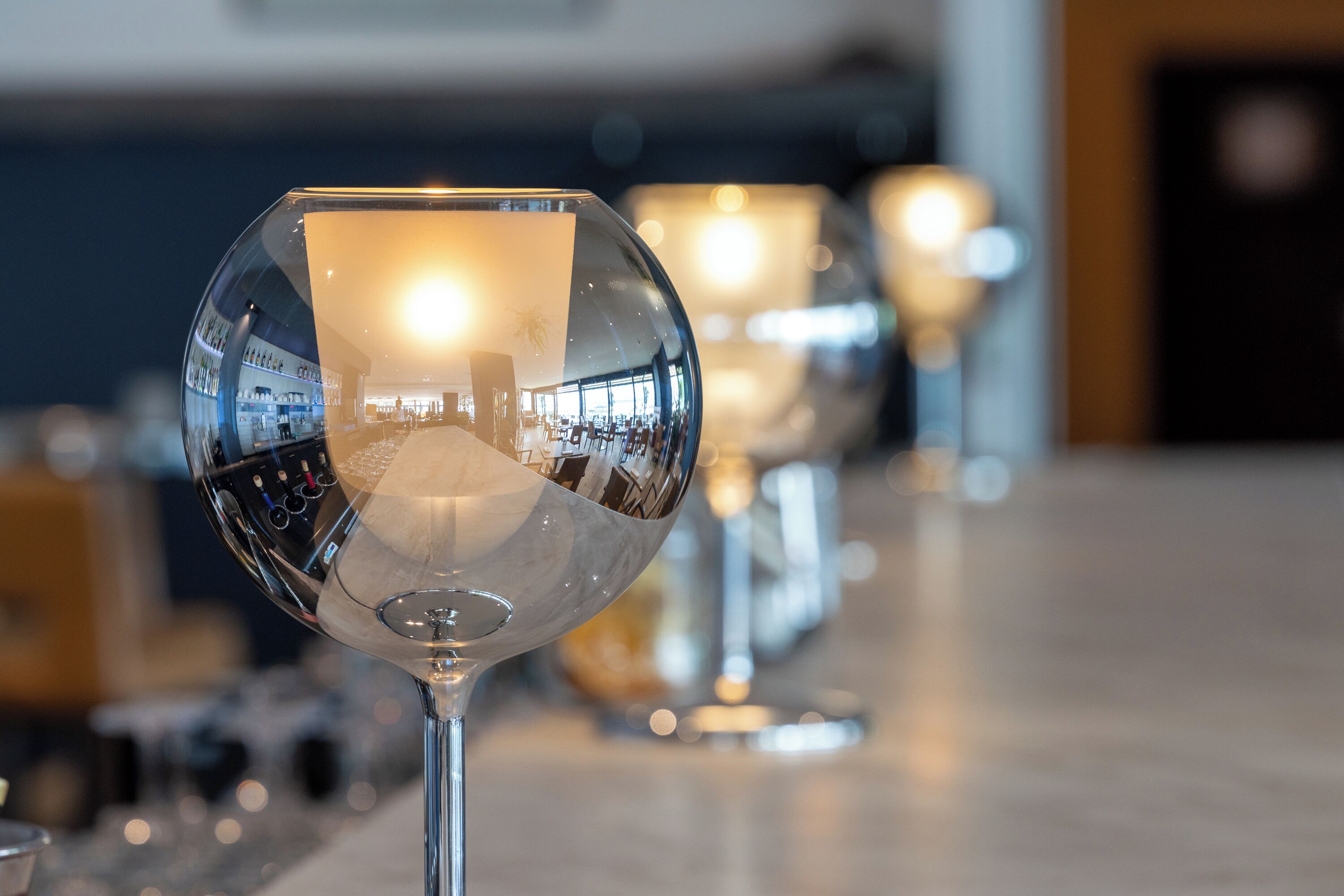 A clear view of a wine glass with a reflection of a bar area, including shelves and lighting.