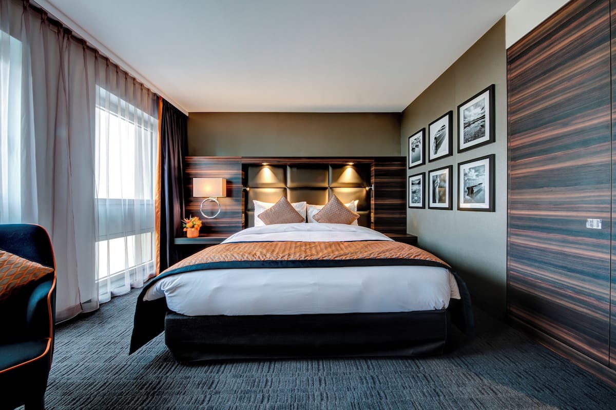 A hotel room with a large bed, a bedside table with a lamp, a chair, and a view of the city through the window.