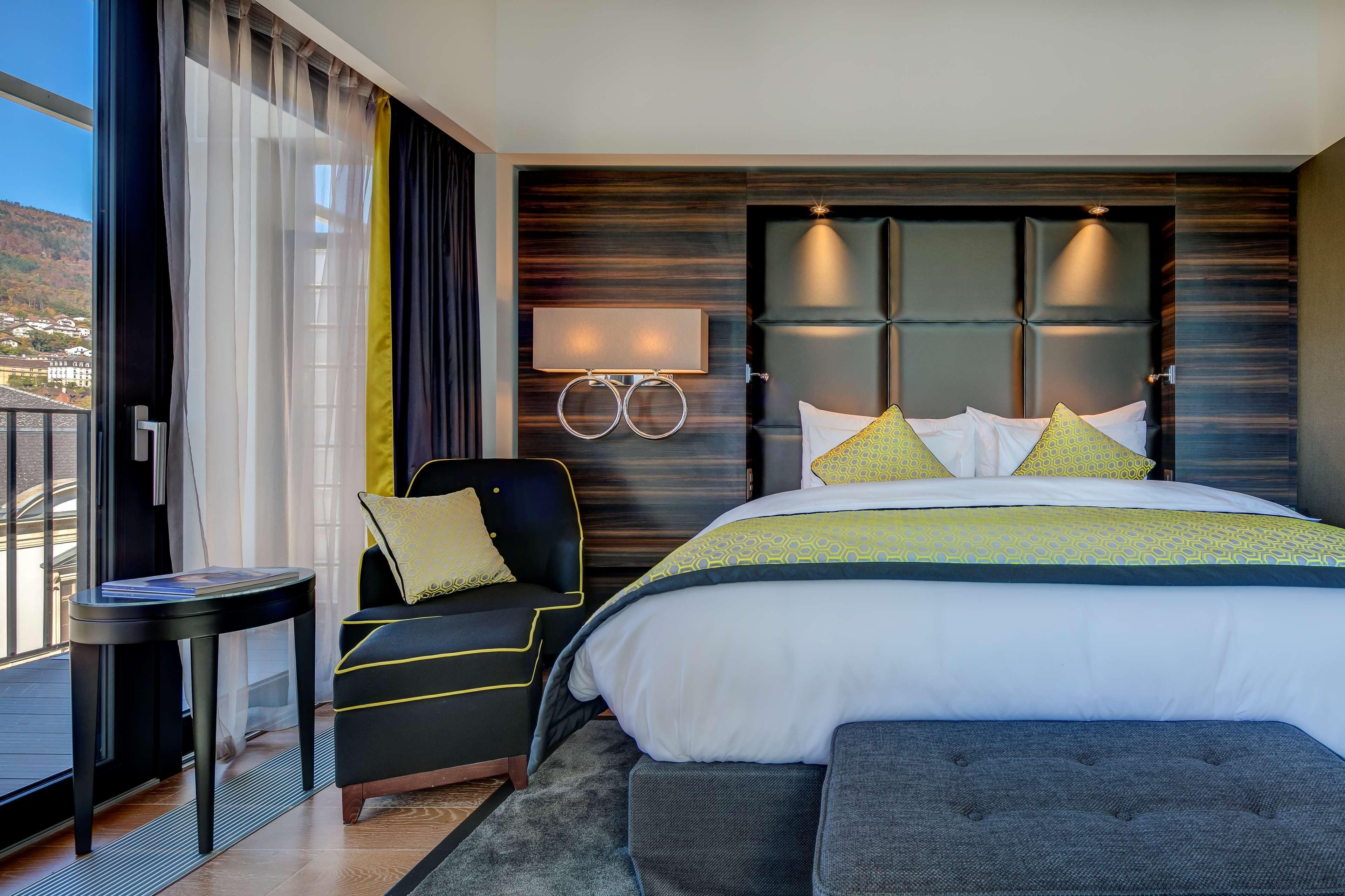 A modern hotel room with a large bed, a bedside table, a chair, and a view of the outside.