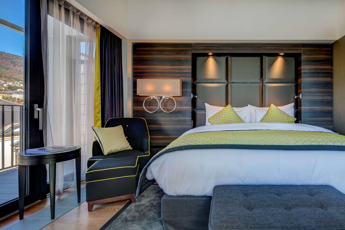 A modern hotel room with a large bed, a bedside table, a chair, and a view of the outside.