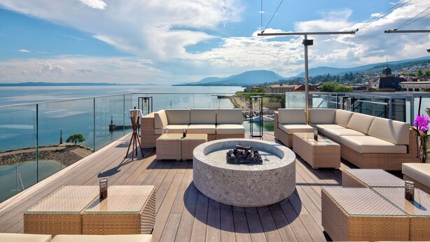 A rooftop terrace with a stone fire pit, cushioned seating, and a glass balustrade overlooking a body of water and distant mountains.
