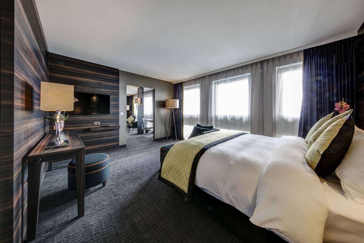 A modern hotel room with a large bed, a desk, a flat-screen TV, and a seating area with a chair and a small table.